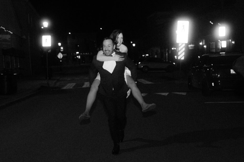 black and white photo of groom giving bride piggyback ride after wedding